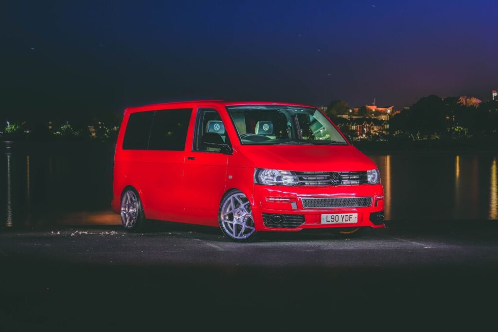 Red Volkswagen van parked by a river at night with city lights in the background.