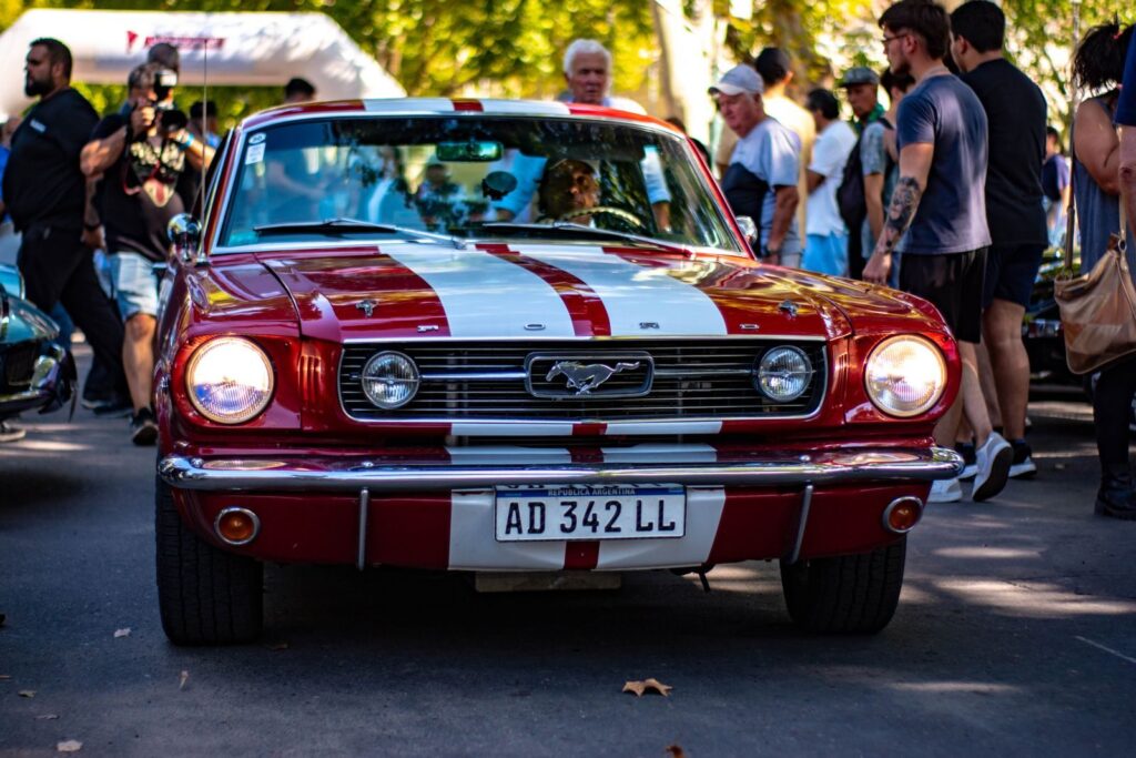 Red classic Ford Mustang showcased at an outdoor car exhibition with crowd.