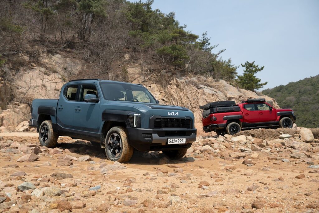 Two modern pickup trucks, Kia Tasman, parked on rocky terrain during a sunny day.