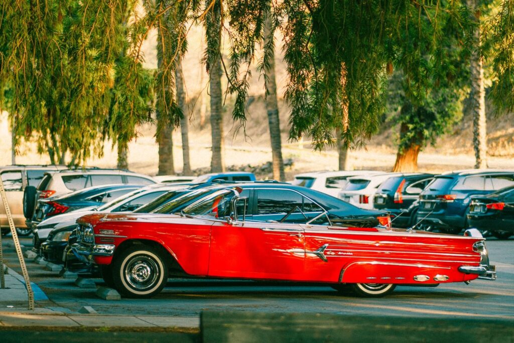 A vibrant red classic car parked among modern vehicles under tall trees in California.