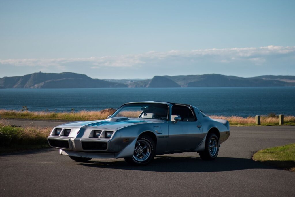 Old luxury retro sports car parked on even asphalt road near shoreline of big lake and mountain range under light blue sky