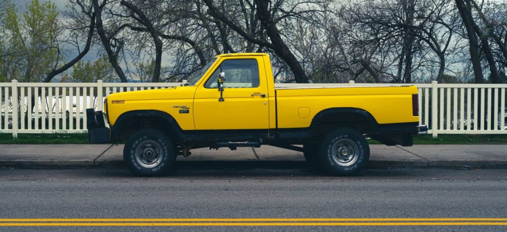 Side view of a parked yellow pickup truck on a city street with trees and a fence in the background.
