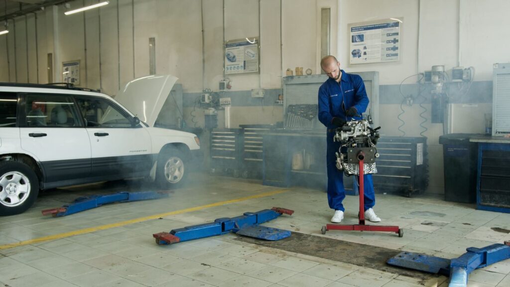Mechanic in blue overalls working on a car engine in an auto repair shop with a white car.