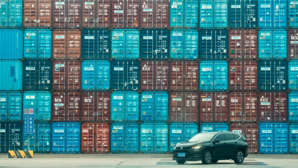 A car is parked in front of a large stack of shipping containers