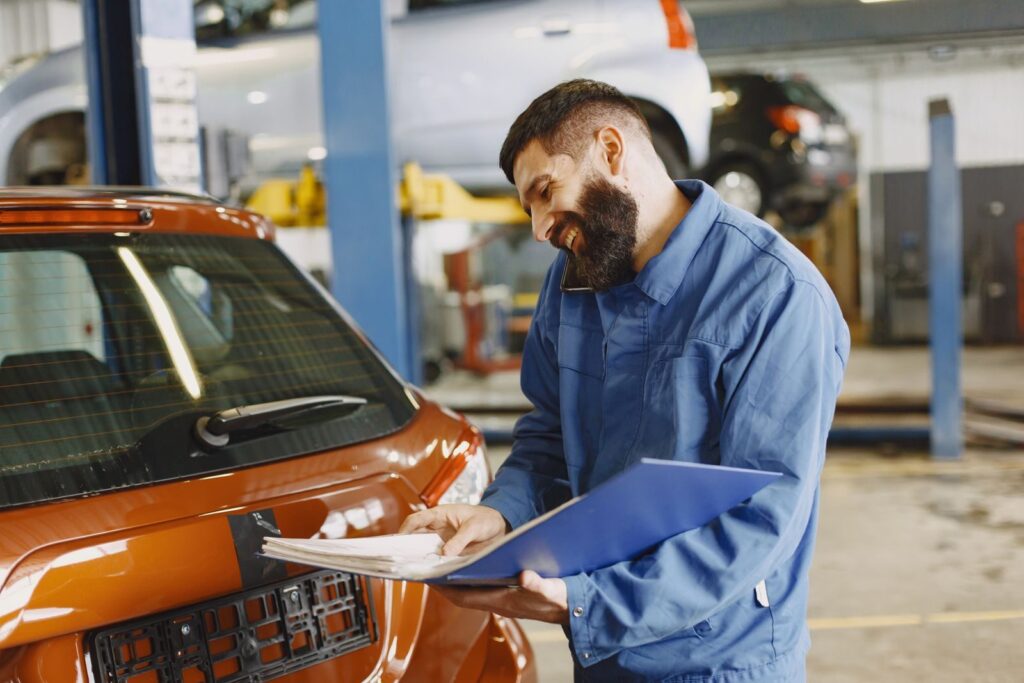 Smiling auto mechanic inspects a car in a service garage, wearing coveralls and holding documents.