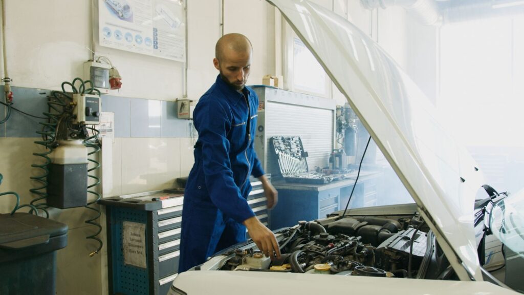 Mechanic in blue uniform inspecting a car engine in a workshop.